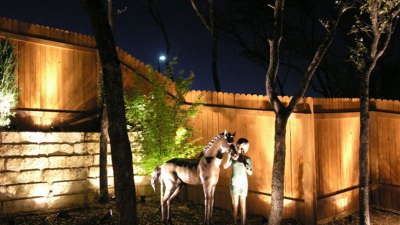 Landscape lighting has illuminated a garden with a statue in it, bordered by a wooden fence